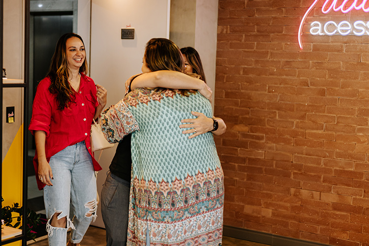 Na porta de entrada da sala da Tudo é Acessibilidade, 3 mulheres se cumprimentam enquanto sorriem. Duas delas se abraçam calorosamente.