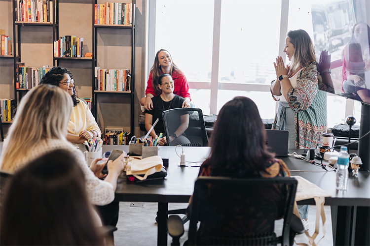 Em volta de uma mesa, algumas mulheres prestam atenção na Ale Miliatti, enquanto ela apresenta algo para todas. Ao fundo, uma estante de livros e a claridade em uma das janelas.