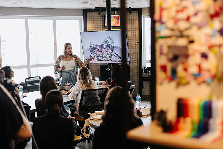 A câmera está posicionada no fundo da sala e é possível ver a Ale Miliatti ao fundo apresentando material da aula enquanto as demais mulheres estão sentadas em volta da mesa prestando atenção.